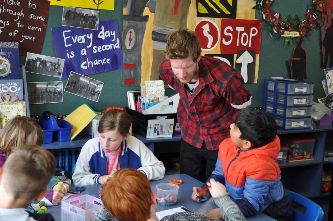 A teacher aide listens to a group of students engaged in a learning task.