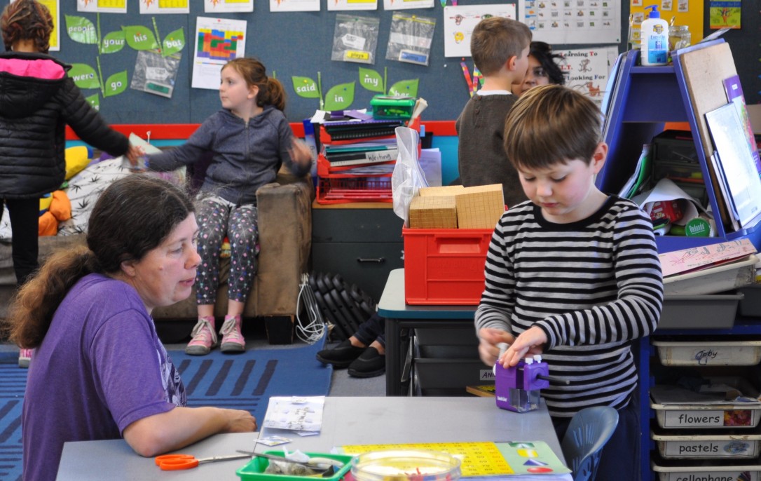 A teacher aide observes a student sharpening a pencil.
